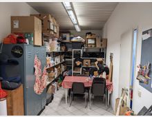 Ein kleiner Lager- und Aufenthaltsraum mit Regalen voller Kartons, Ordner und anderen Gegenständen, sowie einem Tisch mit rot-weiß-karierter Tischdecke und mehreren Stühlen. An der Wand steht ein grauer Spind, und die Decke ist mit funktionalen Deckenleuchten ausgestattet.