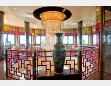 Innenraum eines asiatischen Restaurants mit opulenter Dekoration, darunter ein großer Kristallleuchter und eine bemalte Keramikvase im Vordergrund. Die roten, geometrisch verzierten Geländer und die bunten Fensterrahmen ergänzen das traditionelle asiatische Design.