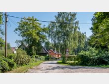 Eine ländliche Straße mit unbefestigtem Weg, umgeben von üppigem Grün und Bäumen. Im Hintergrund stehen Häuser mit Ziegeldächern, und am rechten Straßenrand befinden sich Verkehrsschilder, darunter ein Schild mit Geschwindigkeitsbegrenzung auf 5 km/h.