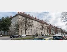 Mehrstöckiges Wohngebäude in heller Putzfassade mit roten Ziegeldächern und Schornsteinen. Vor dem Gebäude stehen Bäume, teils blühend, sowie eine geparkte Reihe von Autos entlang der Straße.