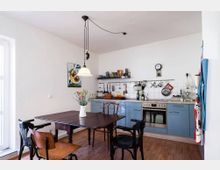 Wohnküche mit hellem Holzboden, weißen Wänden und einer Küchenzeile mit blauen Schränken. Ein Esstisch mit Stühlen steht im Vordergrund, während die Arbeitsfläche mit Küchengeräten, einem Regal, einer Wanduhr und Hängeregalen an einer weißen Fliesenwand im Hintergrund sichtbar ist.