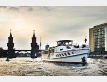 Ein Fluss mit einem weißen Passagierschiff im Vordergrund, im Hintergrund die Oberbaumbrücke mit ihren markanten Türmen und die Skyline von Berlin, einschließlich des Berliner Fernsehturms.
