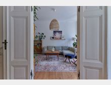 Blick durch eine offene weiße Doppeltür in ein helles Wohnzimmer mit Holzboden. Im Raum befinden sich eine graue Eckcouch, ein Holztisch, ein orientalischer Teppich, ein Sideboard mit Pflanzen und Deko sowie eine geflochtene Hängelampe an der Decke.