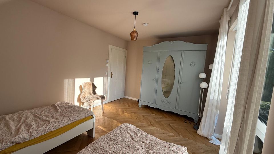 Schlafzimmer mit hellen Wänden, zwei Einzelbetten mit gemusterter Bettwäsche, einem großen, pastellgrünen Kleiderschrank mit ovalem Spiegel und einem Stuhl mit Fellauflage. Der Raum hat einen Holzboden im Fischgrätmuster und große Fenster mit transparenten Vorhängen, die viel Tageslicht einlassen.