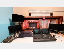 Ein Regieraum mit mehreren Monitoren, Lichtsteuerpulten und Mischpulten, der auf eine kleine Bühne mit geschlossenen Vorhängen und Theaterbeleuchtung blickt. Im Hintergrund ist ein Holzelementrahmen um das Fenster und Dokumente an der Wand sichtbar.