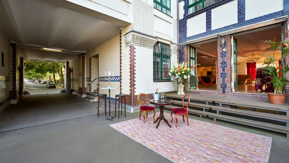 Durchfahrt und Eingangsbereich eines Gebäudes mit blau-weißer Klinkerfassade und großen grünen Sprossenfenstern, rechts mit breiter Treppe zu einem offenen Saal. Im Vordergrund steht eine kleine Sitzgruppe auf einem Teppich sowie Stehtische; im Hintergrund führt die Passage zur baumgesäumten Straße.