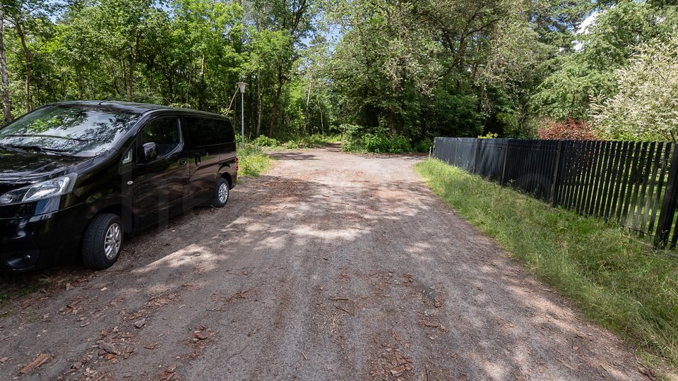 Unbefestigter Waldweg mit breiter, sandiger Fahrspur, umgeben von dichtem Grün und hohen Bäumen. Rechts verläuft ein schwarzer Lattenzaun entlang des Weges, im Hintergrund steht eine einzelne Straßenlaterne.