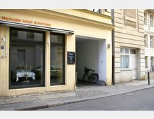 Fassade eines gelb-beigen Gebäudes mit großen Schaufenstern, hinter denen ein Tisch mit weißer Tischdecke steht. Daneben ein Durchgang mit abgestelltem Fahrrad, rechts weitere Hauseingänge und gepflasterter Gehweg.