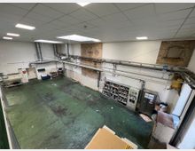 Großer, industrieller Innenraum mit grünem, abgenutztem Boden, weißer Decke mit eingebauten Leuchten und sichtbaren Lüftungsrohren an den Wänden. An der rechten Wand befinden sich Regale mit Farbdosen und ein Schrank, während die Wände teils unverputztes Mauerwerk zeigen.