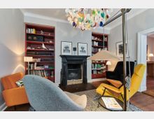 Gemütliches Wohnzimmer mit Kamin in der Mitte, flankiert von zwei roten Bücherregalen. Farbige Designerstühle, darunter ein grauer, ein orangefarbener und ein gelber Sessel, sowie eine auffällige, mehrfarbige Deckenleuchte dominieren den Raum.