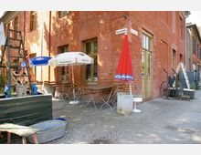 Außenbereich eines Restaurants oder Cafés in einem Backsteingebäude mit Holztüren und Fenstern. Vor dem Gebäude stehen Metalltische und -stühle, Sonnenschirme sowie Dekorationen wie ein alter Wagen und ein Metallgestell mit Töpfen.