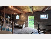 Rustikaler Innenraum eines umgebauten Scheunen- oder Stallgebäudes mit sichtbaren Holzbalken, Ziegelboden und verputzten Steinwänden. Eine offene Tür führt in einen grünen Garten; kleine Sprossenfenster und eine Holztreppe prägen den Raum.