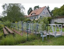 Ein Wohnhaus mit rotem Ziegeldach und grünen Fensterläden steht in einer grünen Umgebung mit Bäumen und Büschen. Im Vordergrund befindet sich ein Zaun, dahinter eine Metallkonstruktion mit Holzstegen, sowie Kunststoffstühle auf einer Wiese.