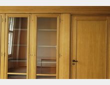 Holzschrankwand mit Glastüren, die Einblick in leere Regalböden gewähren, und einer geschlossenen Holztür mit schwarzem Türgriff. Im Hintergrund sind Fenster und ein Tisch erkennbar.