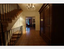 Ein Eingangsbereich mit Fliesenboden, einer Holztreppe mit schwarzem Geländer auf der linken Seite und einem großen, antiken Holzschrank auf der rechten Seite. Im Hintergrund ist ein angrenzender Raum mit Sitzmöbeln und Fenstern zu sehen, ein Kronleuchter hängt von der Decke.