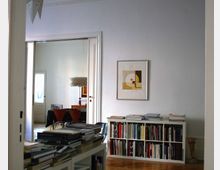 Innenraum einer Wohnung mit einem Durchgang zu einem weiteren Raum. Im Vordergrund ein Bücherregal mit Büchern und Ordnern, darüber gestapelte Bücher; an der Wand ein gerahmtes Bild. Im zweiten Raum ein Esstisch mit Stühlen und ein hängender Lampenschirm.