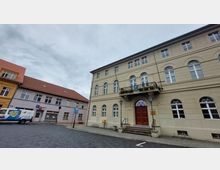 Historisches Gebäude mit hellbeiger Fassade, symmetrischen Rundbogenfenstern und einem Eingang mit einem roten Holztor, darüber ein Balkon mit Blumenschmuck. Rechts und links stehen Straßenlaternen, im Hintergrund befinden sich weitere Gebäude mit roten Ziegeldächern.