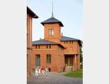 Backsteingebäude eines Standesamts mit einem Turm und dunkelgrauem Dach; im Vordergrund eine gepflasterte Fläche und eine Rasenfläche, auf der vier Personen spazieren.