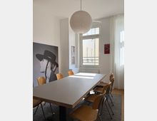 Heller Besprechungsraum mit langem rechteckigem Tisch und mehreren Holzstühlen auf einem Teppich. Große Fenster mit weißen Vorhängen, runde Papier-Hängelampe und Wandkunst an den weißen Wänden.