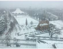 Blick auf eine verschneite Stadtlandschaft mit einer historischen Backsteinruine im Vordergrund, einem Schneefeld und dem markanten Dach eines Veranstaltungsgebäudes (Tempodrom) im Hintergrund. Umgebend sind vereinzelt Bäume, parkende Autos und moderne Gebäude bei stark bewölktem Winterwetter.