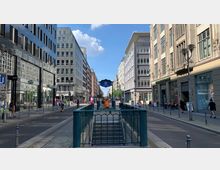 Straßenszene in einer urbanen Umgebung mit mehrstöckigen Gebäuden zu beiden Seiten und einem Eingangsbereich zur U-Bahn-Station "Stadtmitte" in der Mitte des Bildes. Menschen gehen oder fahren auf der Straße, während Geschäfte, Café-Terrassen und Verkehrszeichen die Umgebung prägen.