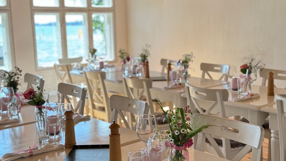 Ein gemütlich eingerichteter Speiseraum mit hellen Holztischen und -stühlen, dekoriert mit Blumenvasen, Servietten und Gläsern; ein Holzbalken an der Decke und große Fenster mit Blick auf Wasser im Hintergrund.
