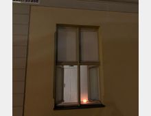 Außenansicht eines gelb gestrichenen Gebäudes mit einem geöffneten Fenster mit vier geteilten Scheiben. Im Inneren ist eine weiße Tür und eine eingeschaltete Kugellampe sichtbar, die warmes Licht abgibt.