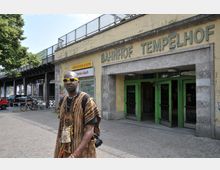 Eingang des Bahnhofs Tempelhof in Berlin mit einem gelben Schild und grünen Türen in einem älteren Gebäude aus Stein. Im Vordergrund ist ein Mann mit einem traditionell gemusterten Oberteil und einer Umhängetasche zu sehen, im Hintergrund Fahrräder und parkende Autos unter einer Hochbahnbrücke.