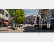 Bahnhofsplatz vor dem Gebäude des Brandenburg Hauptbahnhofs mit Uhr an der Fassade, umgeben von modernen Gebäuden, Bäumen und Geschäften mit Sonnenschirmen. Menschen spazieren durch den Platz, während Fahrräder an Ständern abgestellt sind.
