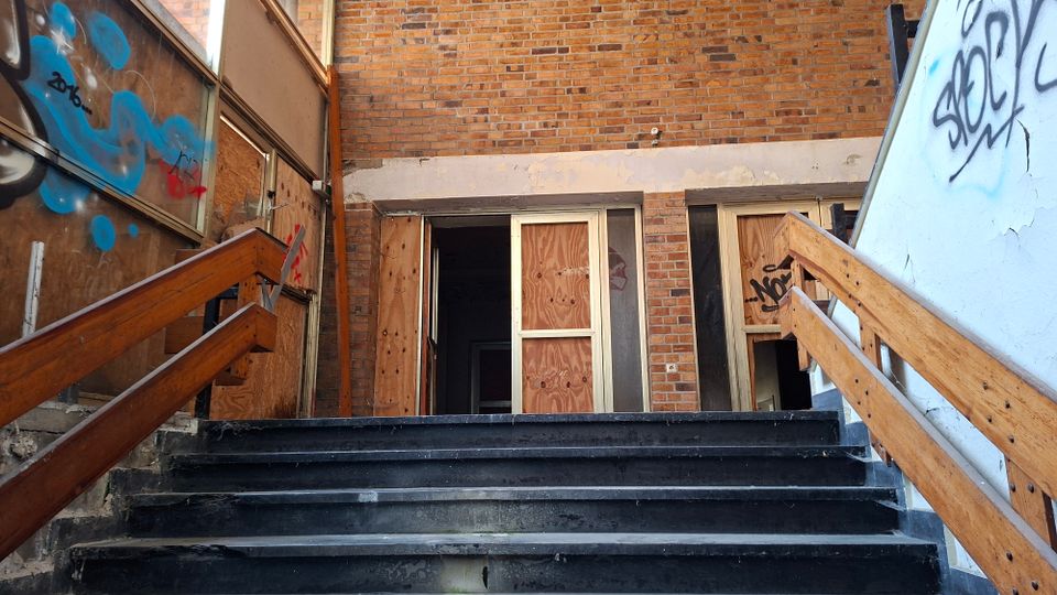 Außentreppe mit dunklen Stufen und Holzgeländern führt zu einem Eingang in einem Backsteingebäude. Fenster und Türen sind mit Sperrholzplatten vernagelt, die Wände zeigen Graffiti und teils beschädigte Oberflächen.