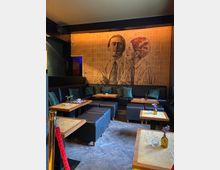 Eine stilvolle Lounge mit dunklen Ledersitzbänken und Hockern, quadratischen Holztischen mit marmorierter Oberfläche und kleinen Blumenvasen. An der Wand hängt ein großformatiges Wandbild, das zwei Personen in Anzügen zeigt, beleuchtet von warmem Licht.