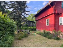 Holzhaus in roter Farbe mit weißen Fenstern, umgeben von einem Gartenbereich mit niedrigem Gras, Büschen und einem hohen Nadelbaum. Im Hintergrund ist ein weiteres modernes Gebäude und dichter Bewuchs sichtbar.