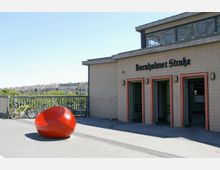 Außenansicht des S-Bahnhofs Bornholmer Straße mit hellbeiger Fassade, drei Eingängen mit roten Rahmen und schwarzer Schrift über der Tür. Im Vordergrund steht eine auffällige rote Sitzskulptur, daneben sind Fahrräder an einem Zaun angelehnt. Im Hintergrund sind Wohnhäuser und Bäume zu sehen.