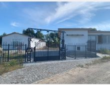 Eingezäuntes Grundstück mit gepflastertem Weg und zwei weißen einstöckigen Gebäuden, eines mit Schiebetür, das andere mit Vordach und Fenstern. Im Vordergrund ein verziertes schwarzes Metalltor mit angrenzendem Zaun, umgeben von niedriger Vegetation und blauem Himmel im Hintergrund.