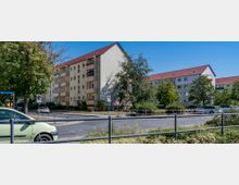 Mehrstöckige Wohngebäude mit hellgelben Fassaden und roten Satteldächern, umgeben von Bäumen und einer Straße mit parkenden Autos im Vordergrund. Ein Straßenschild mit der Aufschrift „Augusta-Bebel-Straße“ und eine Verkehrsinsel sind sichtbar.