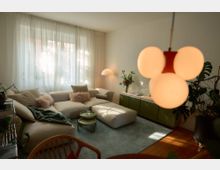 Wohnzimmer mit hellbeiger Eckcouch und passendem Hocker auf einem hellblauen Teppich. Im Hintergrund befinden sich ein Sideboard in Olivgrün, eine Stehlampe und Zimmerpflanzen vor einem Fenster mit weißen Vorhängen, während eine moderne, kugelförmige Deckenleuchte im Vordergrund hängt.