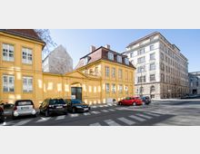 Gelbes zweistöckiges Gebäude mit Satteldach und großen Fenstern, davor parkende Autos auf der Straße; rechts daneben ein mehrstöckiges, graues Steingebäude mit klassischer Fassade. Klarer Himmel und urbanes Umfeld.