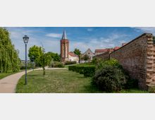 Stadtpark mit Rasenflächen, Gehweg und Laternen, flankiert von einer alten Backsteinmauer rechts und Bäumen links; im Hintergrund ein historischer, runder Kirchturm mit spitzem Dach sowie Häuser mit roten Ziegeldächern.