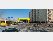 Eingang zum U-Bahnhof Jakob-Kaiser-Platz mit gelbem Überdachungsbau, umgeben von geparkten Fahrrädern. Im Hintergrund sind ein Bus, Verkehrsschilder und ein mehrstöckiges Wohngebäude zu sehen.