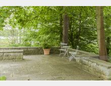 Steinerne Terrasse umgeben von dichtem Grün, mit weißen Metallstühlen, einem runden Tisch und einem großen Pflanzentopf; im Hintergrund stehen Bäume und teils ist Wasser sichtbar.