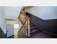 Treppenhaus in einem älteren Gebäude mit dunkelroten Holzstufen und einem gelb lackierten Geländer. Die Wände sind teils dunkelgrau und weiß gestrichen, und eine offene Tür führt in einen angrenzenden Raum.