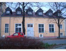 Gelbes Gebäude mit symmetrischen Dachgauben und großen weißen Fenstern, umgeben von kahlen Bäumen; ein rotes Auto ist am Straßenrand geparkt, und ein Metallzaun steht vor dem Gebäude.