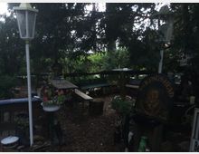 Ein schattiger Garten mit Holztischen, Bänken und dekorativen Elementen, darunter Blumenampeln und eine alte Laterne. Im Vordergrund ist ein Schild mit der Aufschrift "The Kings Head" und ein rustikales Ambiente mit viel Grün und Holz zu erkennen.