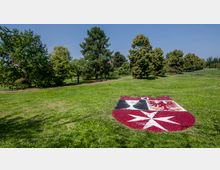 Eine Grünfläche in einem Park mit Bäumen im Hintergrund und einem großen Wappen aus farbigen Blumen im Vordergrund, das auf dem Rasen angelegt ist. Ein Weg führt zwischen den Bäumen hindurch, und einige Bänke stehen im Schatten.