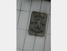 Metallschild mit der Aufschrift "AUFBAU PROGRAMM 1953" und einem Bärenemblem, montiert an einer Wand mit hellgrauen rechteckigen Fliesen.