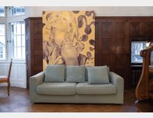 Wohnraum mit einer hellblauen Couch im Vordergrund, darüber ein großes Gemälde mit einer stilisierten Darstellung einer Frau auf gelbem Hintergrund. Der Raum ist durch Holzvertäfelungen, ein weiß gerahmtes Fenster und klassische Möbelstücke gekennzeichnet.