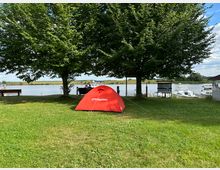 Rote Campingzelt steht auf einer Wiese unter zwei großen Bäumen, im Hintergrund befindet sich ein See mit Booten und ein Uferbereich mit Sitzbänken. Der Himmel ist teils bewölkt, und die Umgebung wirkt ruhig und naturnah.