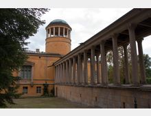 Historisches Gebäude mit gelber Fassade und Rundturm mit grüner Kuppel, daneben ein Säulengang mit klassizistischen, verzierten Säulen; umgeben von Bäumen und einer Rasenfläche.