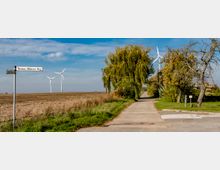 Kreuzung eines asphaltierten Weges mit einem Straßenwegweiser „Thomas-Müntzer-Weg“, umgeben von offenen Feldern, Windrädern in der Ferne und grünen Bäumen entlang des Weges.