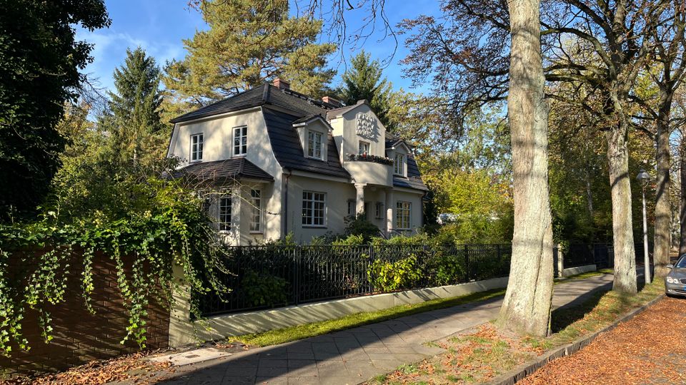 Einfamilienhaus in einer grünen Wohngegend mit hellem Putz, dunklem Mansarddach und vielen Sprossenfenstern, umgeben von Bäumen und Sträuchern hinter einem schmiedeeisernen Zaun. Davor verläuft ein Gehweg entlang einer von großen Bäumen gesäumten Straße mit Herbstlaub am Boden.